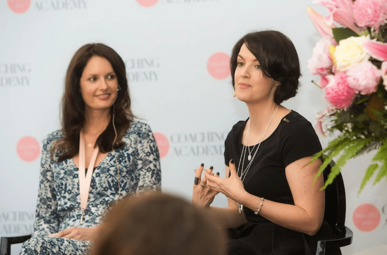 Photo of two women sitting on stage, Beautiful You Coaching Academy banner in background. One woman looks at the other who is speaking.